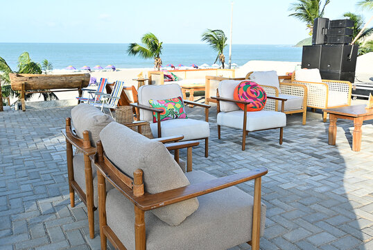 outdoor beach lounge deck with wooden armchairs and colorful patterned cushions, seaside event setup on paved stone terrace with ocean view and palm trees