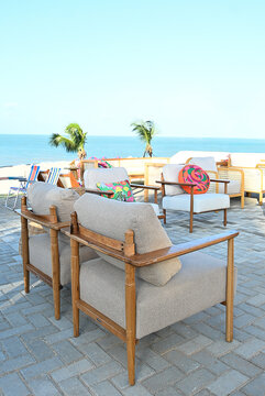 outdoor beach lounge deck with wooden armchairs and colorful patterned cushions, seaside event setup on paved stone terrace with ocean view and palm trees