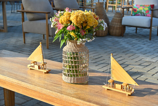 yellow flower bouquet in woven glass vase and miniature wooden sailboat models on coffee table, rustic beach event setup with wicker lounge chair in background