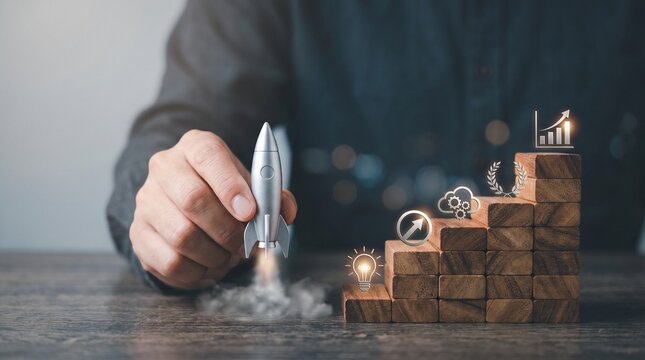 Close up of hand moving silver rocket model upward next to wooden stairs featuring holographic business growth symbols.