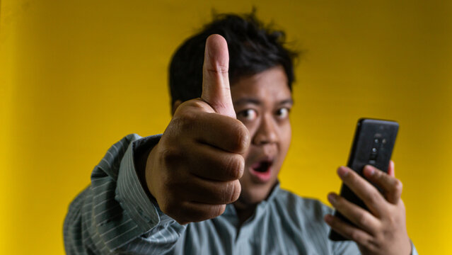 A young Asian male gives a prominent thumbs up gesture towards the camera while holding a modern mobile device, set against a solid yellow background. The hand and thumb are in sharp