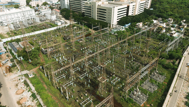 Aerial view of an electrical power station with metal towers, power lines, and transformers surrounded by urban buildings and greenery in India.