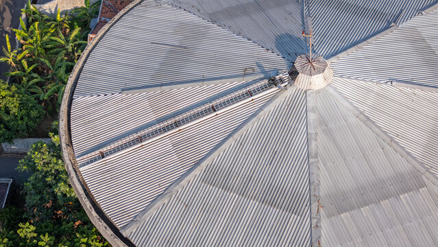 Aerial view of a circular corrugated metal roof with a central ventilation structure surrounded by lush green trees in Klaten, Central Java, Indonesia.