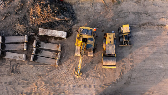 Aerial view of heavy construction machinery including an excavator, a road roller, and a bulldozer parked on a dirt site with concrete blocks in Klaten, Central Java, Indonesia.