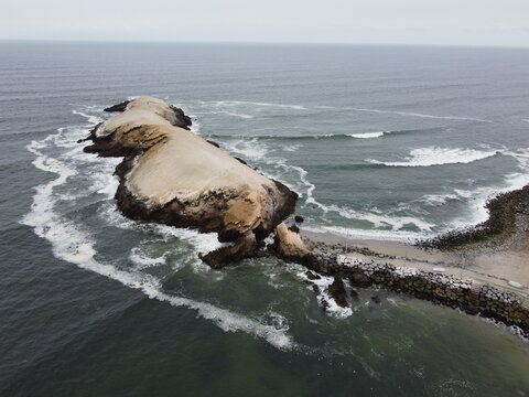 View of the rocky Isla Punta Hermosa surrounded by the Pacific Ocean with waves crashing against the shore and a stone breakwater in Punta Hermosa, Lima Province, Peru.
