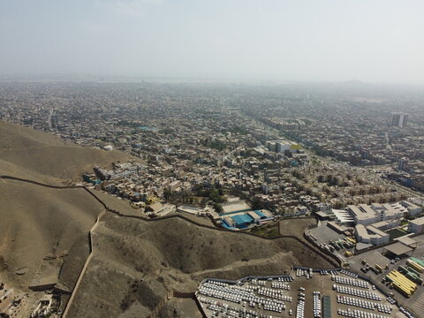 View of Cerro La Milla overlooking the dense urban sprawl of San Martin de Porres with residential rooftops and a large vehicle storage lot in Lima Province, Peru.
