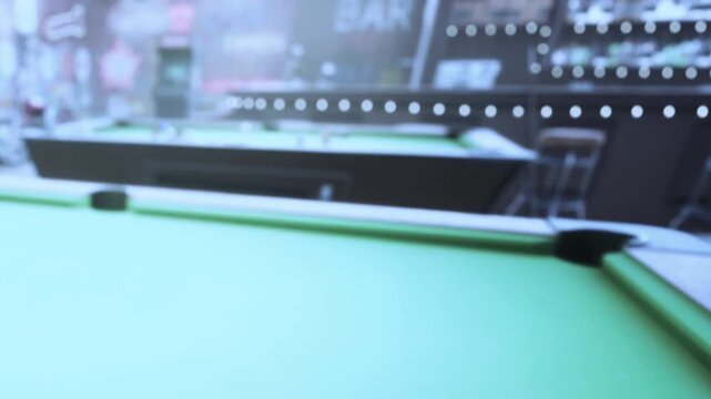 A green felt pool table is in focus while other tables and visitors are in the background. It is evening and there is activity in a bar setting.