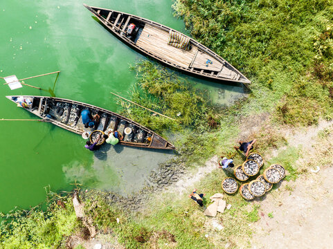 Dhaka, Bangladesh - 01 May 2024: Aerial view of fishermen sorting their catch into baskets on the riverbank with wooden boats moored in the green water.