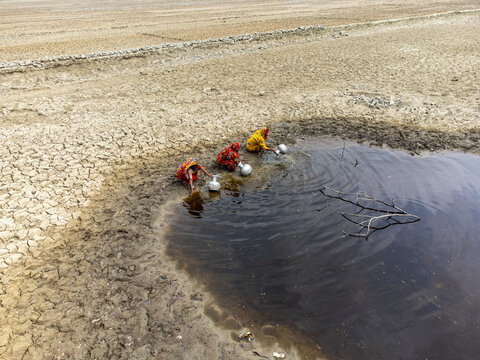 Shyamnagar, Khulna Division, Bangladesh - 25 April 2025: Aerial view of three women in colorful saris collecting water from a small, muddy pool in a dry, cracked landscape.
