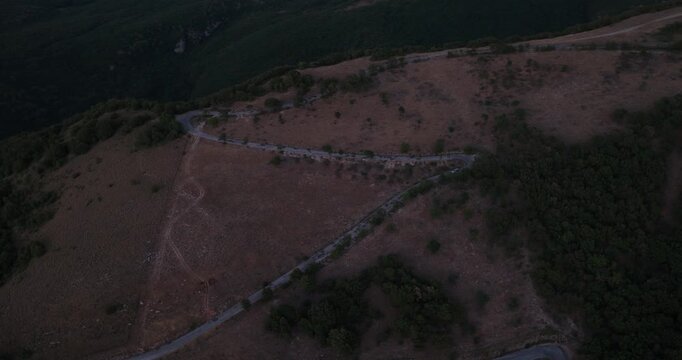 Aerial view of a winding road on the hillside of Monteluco at dusk, featuring sparse trees and dry terrain in Monteluco, Spoleto, Umbria, Italy.