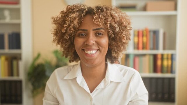 Woman smiling showing teeth in a white shirt seated before a bookshelf with plants and binders in a building, facing camera; calm joy.