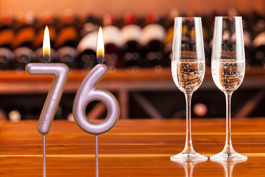 Birthday card, anniversary or celebration of a special day, lit candles forming the number 76 and two champagne glasses; Photo on a background of a bookshelf with blurred wine bottles.