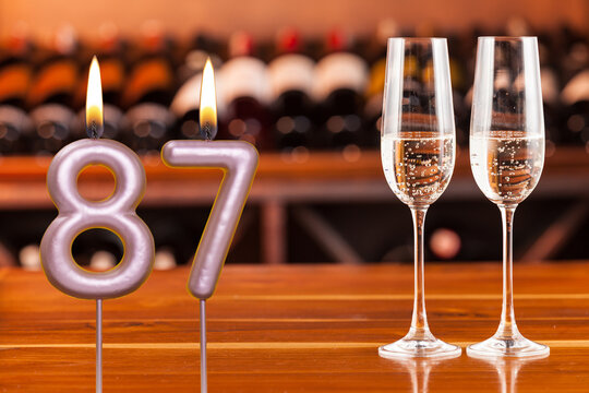 Birthday card, anniversary or celebration of a special day, lit candles forming the number 87 and two champagne glasses; Photo on a background of a bookshelf with blurred wine bottles.