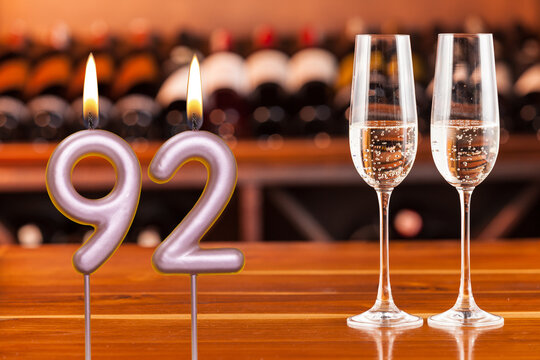 Birthday card, anniversary or celebration of a special day, lit candles forming the number 92 and two champagne glasses; Photo on a background of a bookshelf with blurred wine bottles.