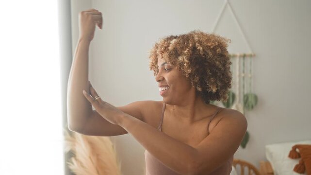 Woman touching her arm and smiling in a bedroom revealing bra strap and natural curls; confidence selfcare.