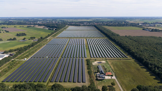 Aerial view of a large solar farm with rows of photovoltaic panels set in a rural landscape with green fields and forest edges in Oude Pekela, Groningen, Netherlands.