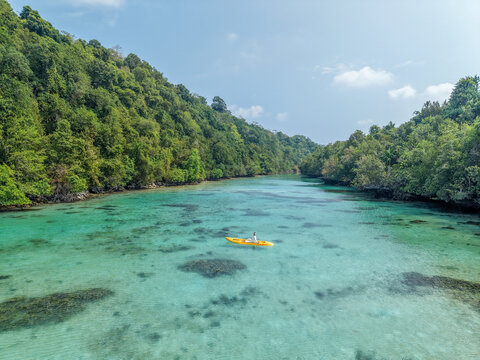 Aerial view of a person paddling a yellow kayak in clear turquoise waters surrounded by lush tropical hills under a bright sky, Indonesia.