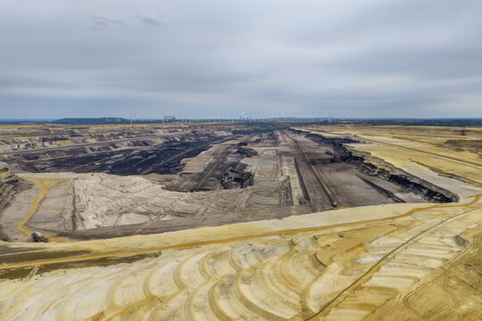 Aerial view of the Garzweiler surface mine showing vast open-pit excavation layers, industrial machinery, and wind turbines on the horizon in Juechen, North Rhine-Westphalia, Germany.
