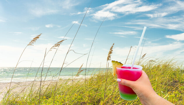 Hand holding a glass with cocktail and straw, Sand Dunes with Grass on Beach with ocean on background