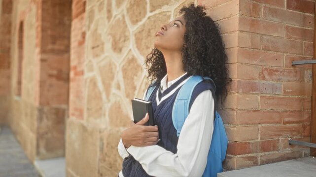 Woman student with backpack holding book, leaning against brick building and looking up; thoughtful reflection.
