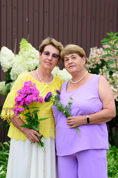 Two senior russian sisters 80 and 75 years old holding flower bouquets standing together in a backyard garden in a summer day in Russia