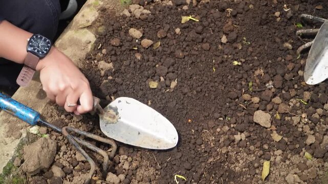 Close-up of hands using trowel to dig into rich soil with gardening tools nearby, capturing earthy texture and preparation for planting in a natural outdoor setting.