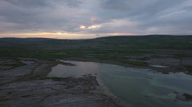 Dramatic aerial footage of the burren region during a cloudy sunset, showcasing a serene lake and expansive rocky landscape.