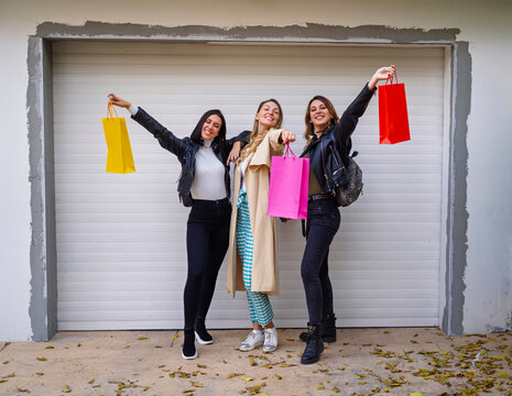 Three smiling young women pose together outdoors while playfully holding up colorful shopping bags, Autumn fashion shopping concept 