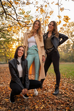 Three smiling fashionable young women pose together outdoors in a park surrounded by golden autumn foliage