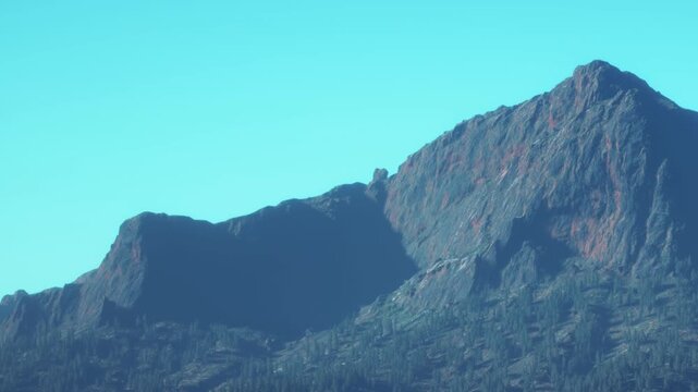 A mountain stands tall against a clear blue sky. The sunlight reveals the rocky surface and the trees below. This scene shows nature in its simple form on a bright day.