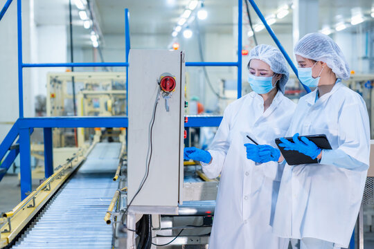 Female quality control inspectors operating control panel in food factory plant, Factory workers in protective uniform and face mask checking production line machine in manufacturing facility
