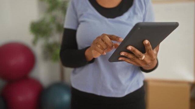 Young african american woman standing in a fitness studio near exercise balls and whiteboard, wearing athletic top, tapping tablet with fingers; focused.