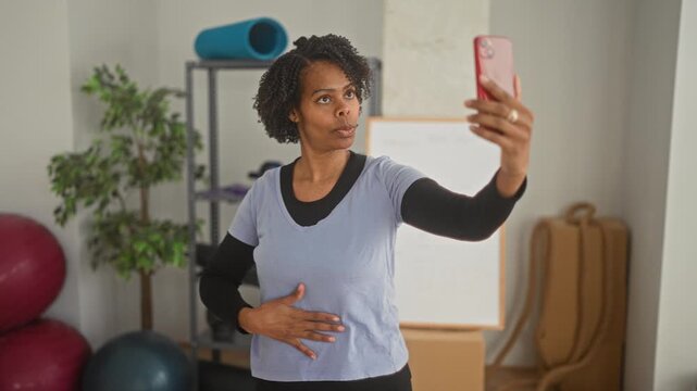 Woman holding smartphone with outstretched arm while gesturing to her stomach during yoga training in a studio; self coaching focus.