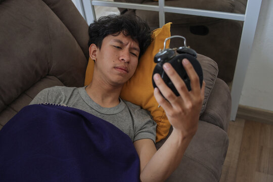 Young Asian Man Looking Annoyingly at an Alarm Clock While Waking up on Brown Sofa at Home