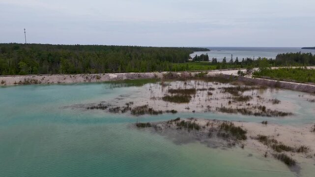 Aerial view crosses a turquoise quarry pond with flooded vegetation pale gravel berms and forest in the distance.