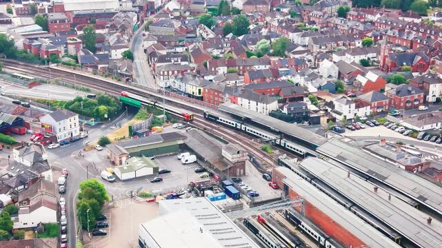 Drone footage of a passenger train passing through an English town's railway station and bridge, with terraced housing, high street shops, and platforms visible below.
