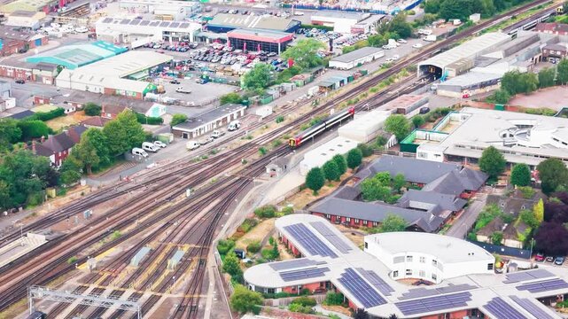 Drone footage of a train approaching a multi-track railway junction, bordered by an industrial estate, car dealership, solar-panelled rooftops, and residential housing.