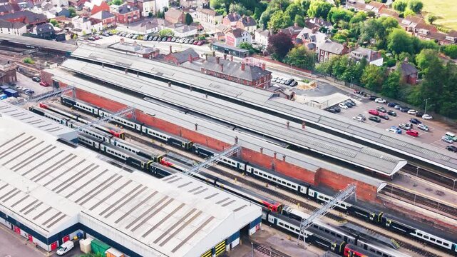Drone footage of a busy railway station showing several trains at platforms, red brick station buildings, covered canopies, a car park, and surrounding residential streets.