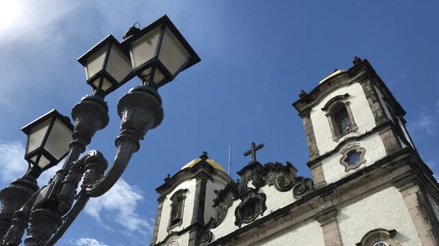 Low-angle view of the historic Church of Nosso Senhor do Bonfim in Salvador, Bahia, showcasing its beautiful colonial architecture against a bright blue sky with wispy clouds and a lamppost