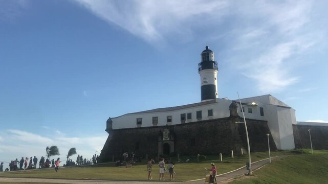 Historic Farol da Barra lighthouse and fort standing against a blue sky with wispy clouds, while tourists enjoy a sunny day on the grassy hill in Salvador, Bahia, a popular landmark