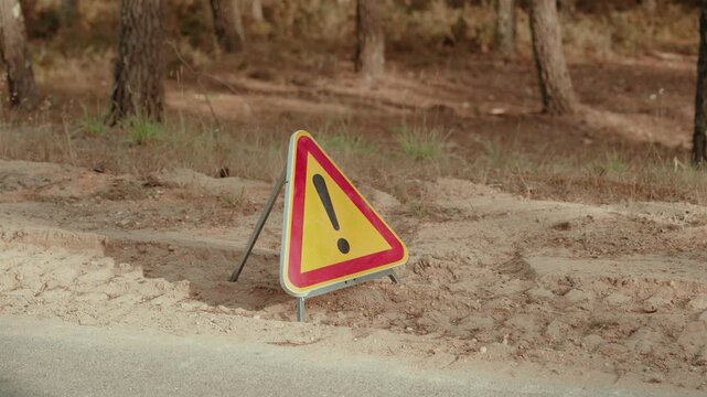 Portable warning triangle sign standing on a sandy roadside beside pine trees