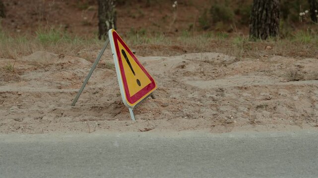 Tilted warning triangle sign on a sandy roadside verge beside pine trees