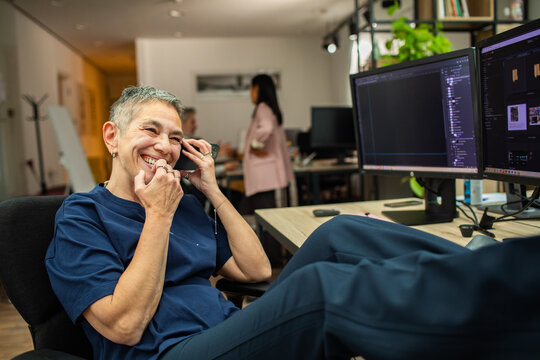 Smiling mature woman on phone at dual monitors in modern office