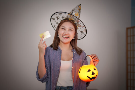 Asian woman wearing witch hat holding credit card and pumpkin lantern for Halloween shopping sale