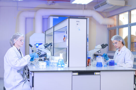 A pair of female lab technicians in white coats, protective eyewear and sterile hairnets holding a tube up across the shared bench in a modern medical research laboratory.