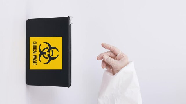 Doctor hand carefully places sharp contaminated medical equipment into wall mounted black biohazard clinical waste container for safe hazard disposal