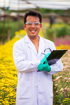 Smiling scientist man holding clipboard in flower nursery, wearing gloves and goggles for horticulture science research among colorful blooming plants