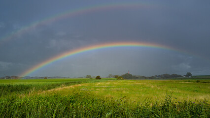 Naklejka premium Rainbow arches across green field. Colorful rainbow follows dramatic weather. Rainbow enhances rural landscape. Brilliant rainbow dominates cloudscape naturally.