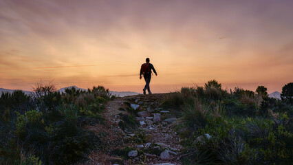 Lone backpacker walking a stony ridge trail into a warm orange and lilac sunset above distant peaks © Kafi