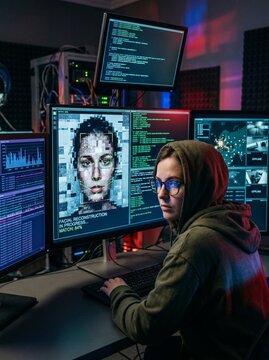 A woman with glasses and a hoodie sits in front of multiple computer screens displaying code and a digital face, suggesting a focus on cybersecurity or data analysis